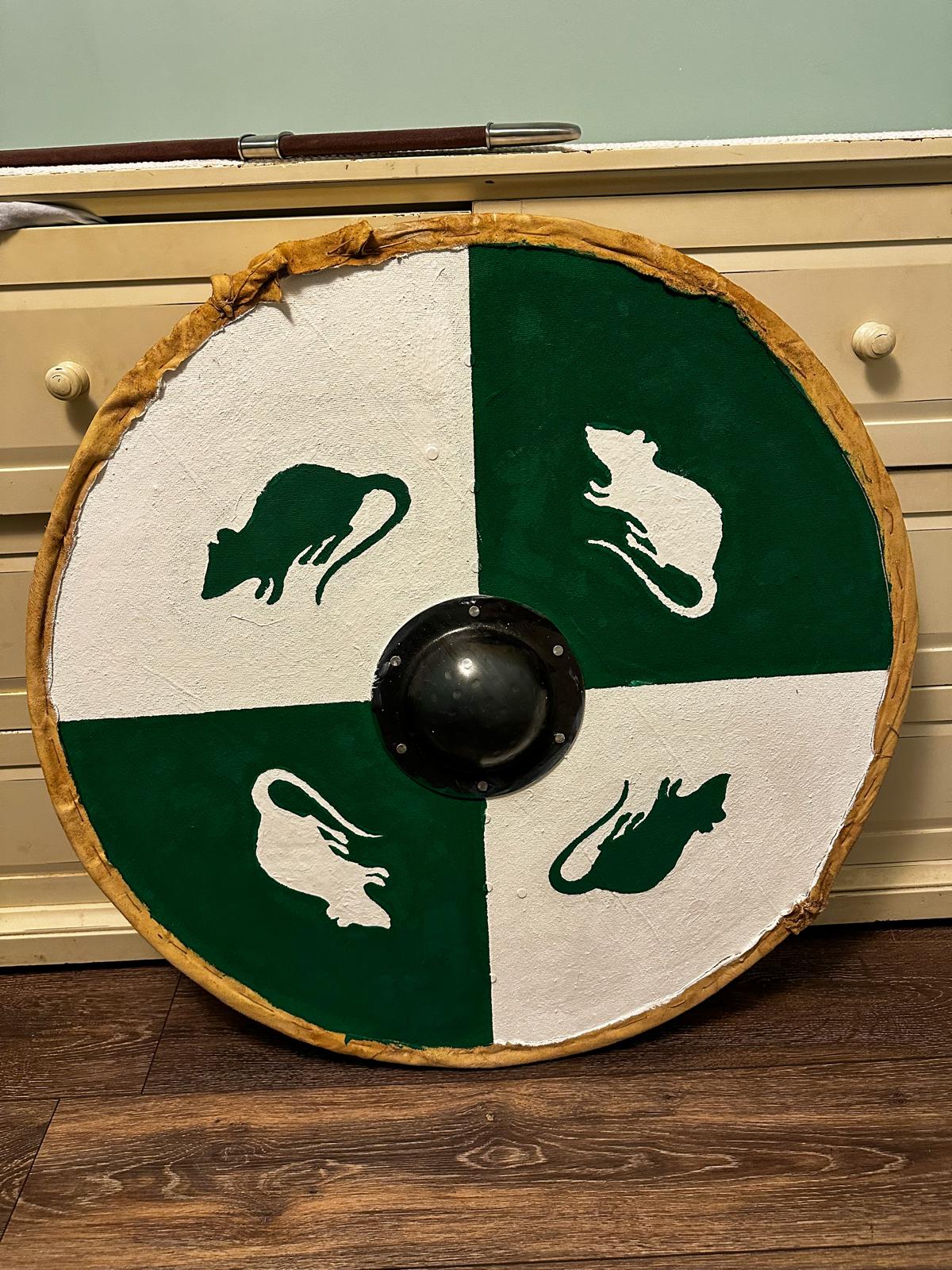 A handmade Viking shield, the paint breaking it into four quadrants, two deep green, two white, each with a single rat of the opposing color emblazoned on it